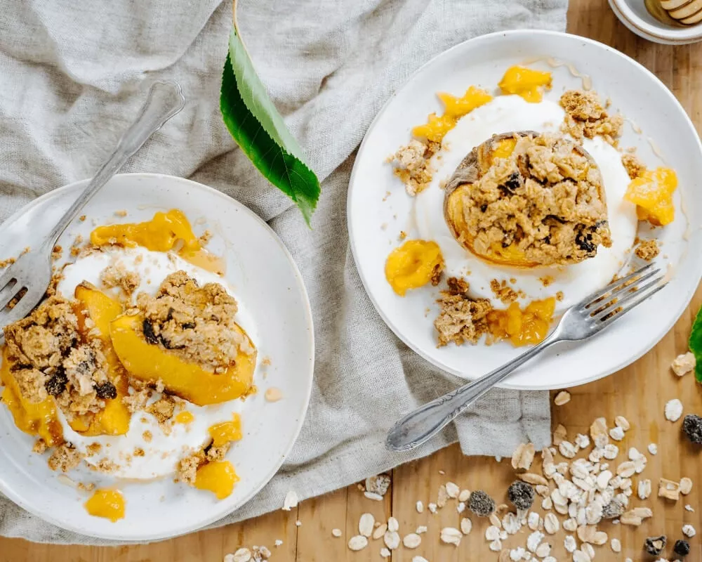 Zwei Teller mit gebackenen Pfirsichen mit Müsli, Joghurt und Mangostücken auf einem Holztisch mit einem Leinentuch.