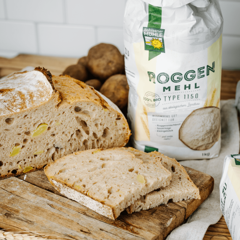 Bohlsener Mühle Roggenmehl Typ 1150 neben einem aufgeschnittenen Brot mit Kartoffelstücken auf einem Holzbrett.