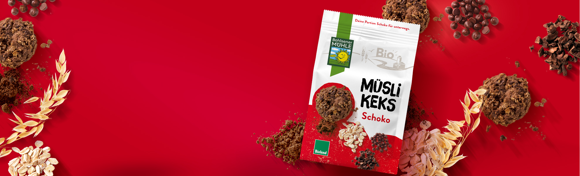 Bohlsener Mühle Müsli Keks Schoko Packung umgeben von Haferflocken und Schokostückchen auf rotem Hintergrund.