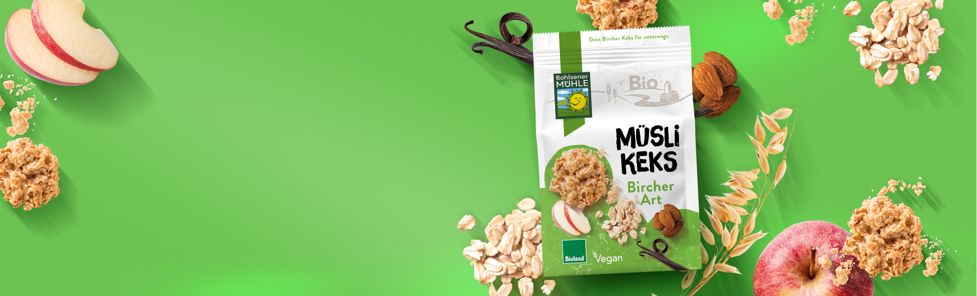 Bohlsener Mühle Müsli Keks Bircher Art umgeben von Äpfeln, Mandeln, Haferflocken und Vanilleschoten auf grünem Grund.