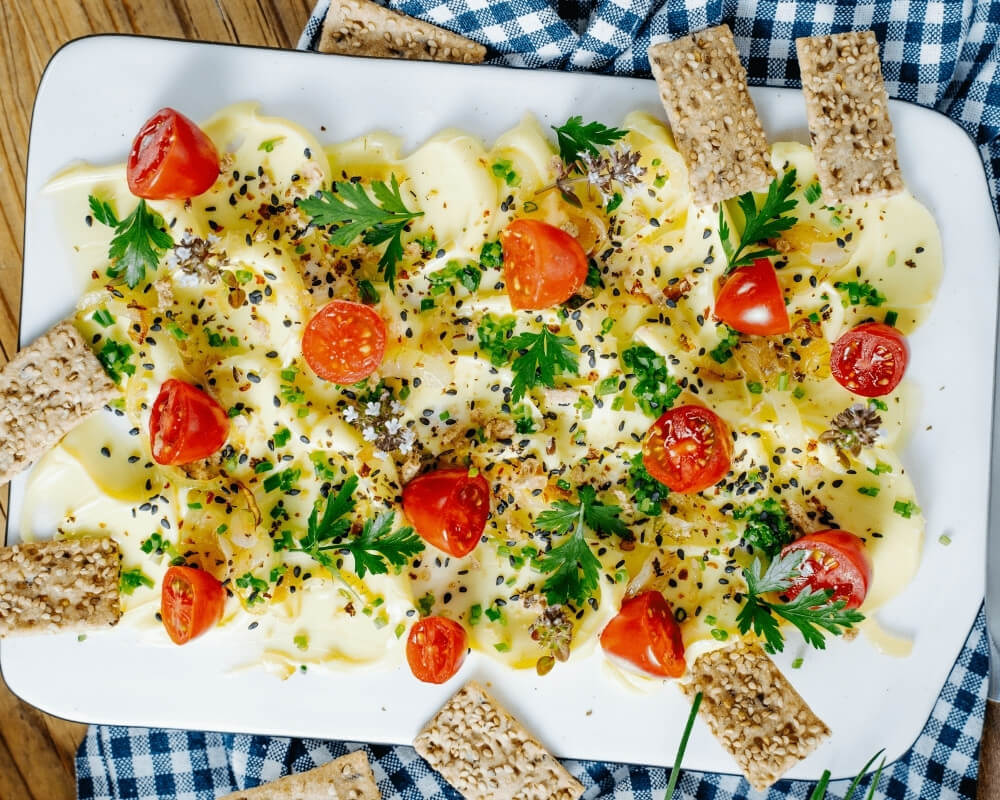 Ein cremiger Aufstrich mit Kirschtomaten, Kräutern und Samen, serviert mit knusprigen Crackern auf einer weißen Platte und einem blau karierten Tuch.