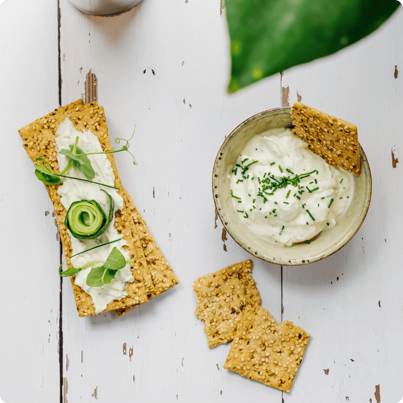 Bohlsener Mühle Crunchy Cracker mit Kräuterquark, Gurke und Schnittlauch auf einem weißen Holztisch serviert.