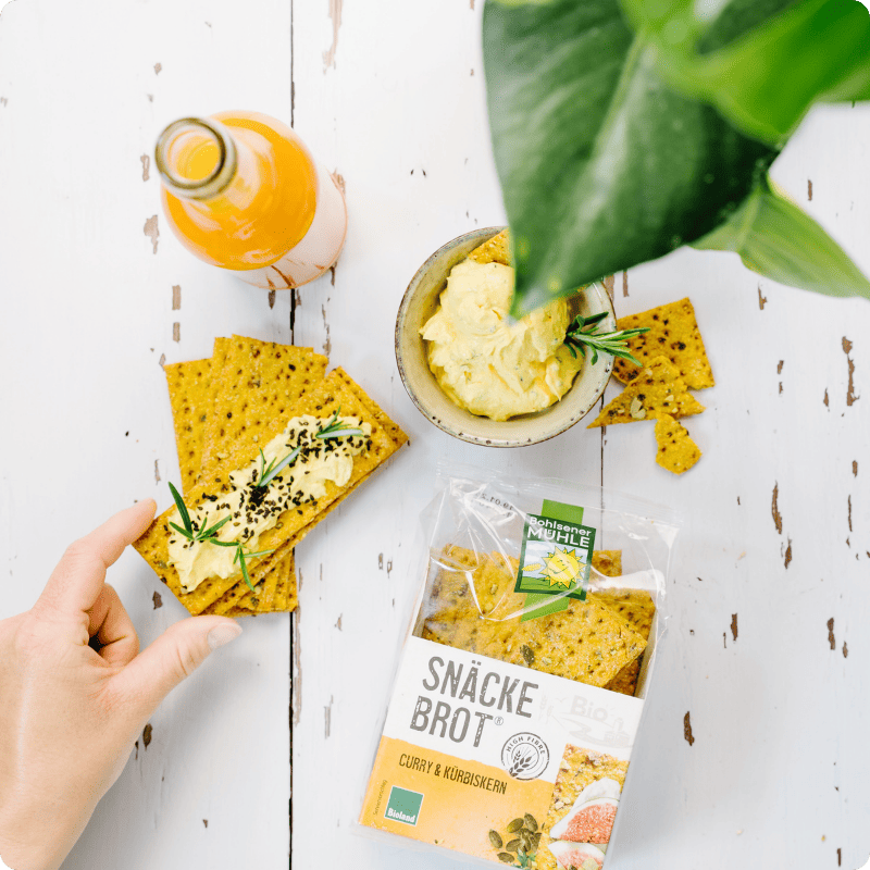Bohlsener Mühle Snäcke Brot Curry & Kürbiskern mit Dip und Getränk auf einem hellen Holztisch angerichtet.