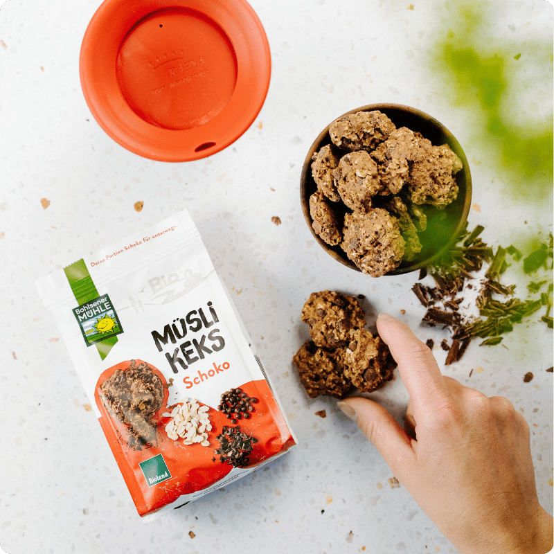 Eine Hand nimmt einen Bohlsener Mühle Müsli Keks Schoko aus einer Schale neben der Produktverpackung.