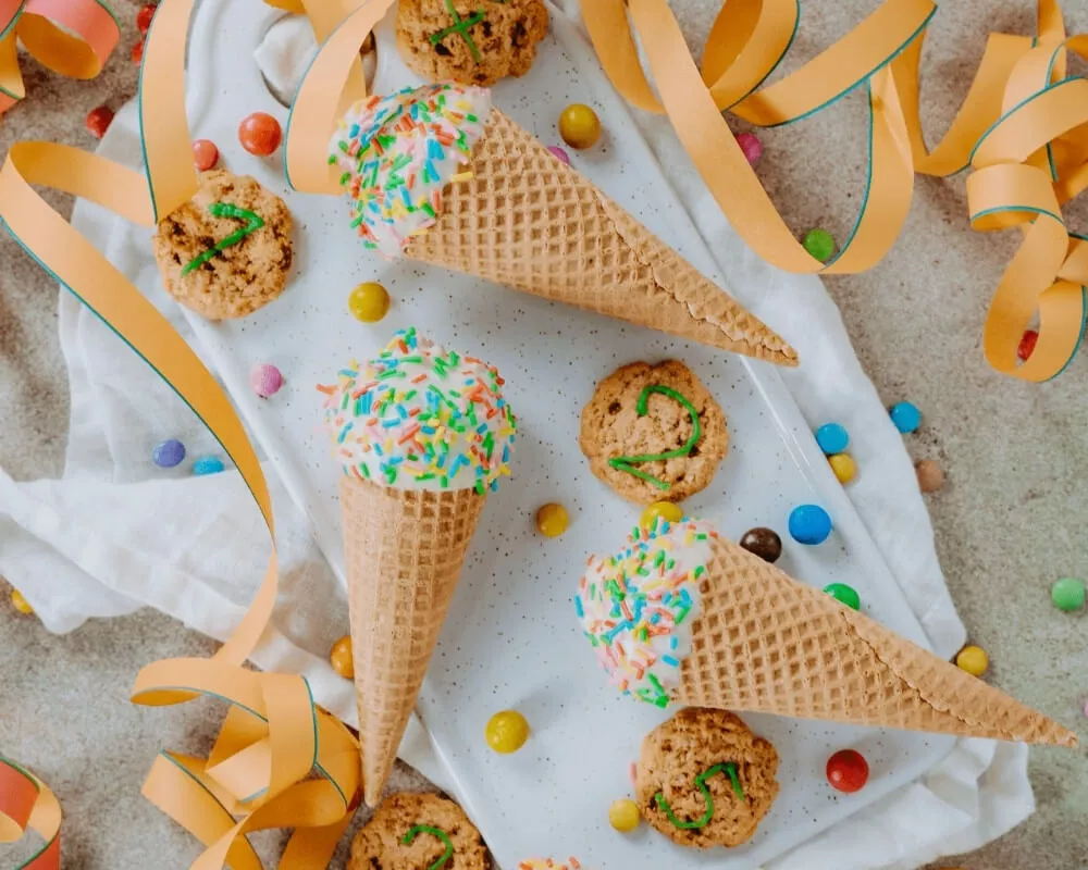 Drei Eistüten mit Streuseln und Keksen auf einem weißen Tablett, umgeben von bunten Bonbons und orangefarbenen Bändern.
