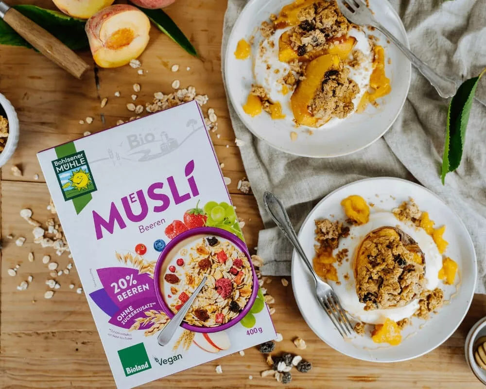 Bohlsener Mühle Bio-Beeren-Müsli-Box neben zwei Tellern gebackener Pfirsiche mit Joghurt und Müsli-Crumble.