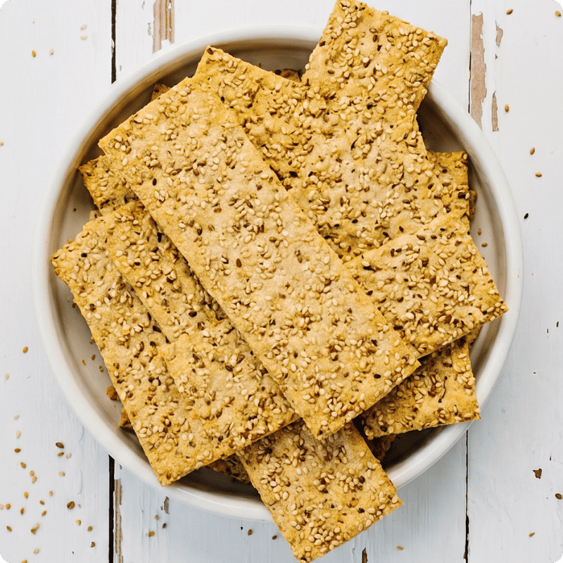 Eine weiße Schale gefüllt mit knusprigen Bohlsener Mühle Sesam-Knäckebrot Stücken auf einem hellen Holztisch.