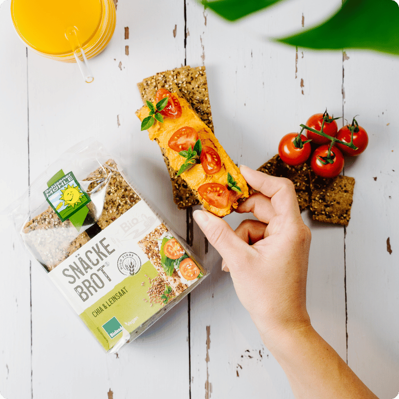 Eine Hand hält ein mit Tomaten belegtes Snäcke Brot Chia & Leinsaat der Marke Bohlsener Mühle auf weißem Holz.