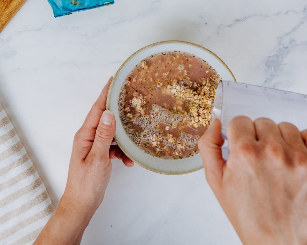 Hände gießen Flüssigkeit in eine Schüssel mit Bohlsener Mühle Cookie Crunchy Müsli auf einem hellen Untergrund.