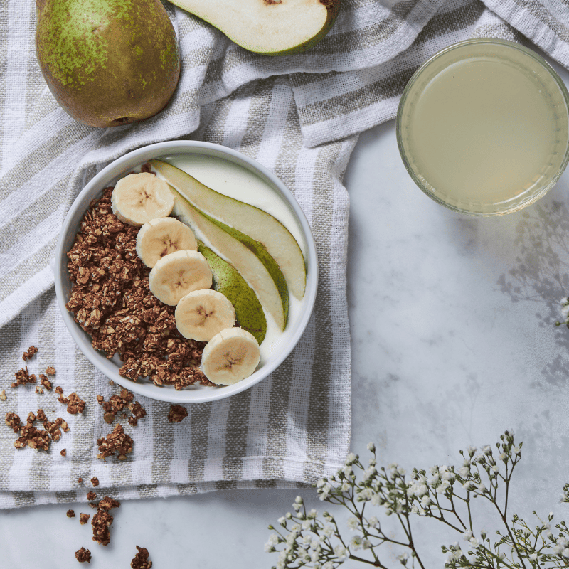 Bohlsener Mühle Cookie Crunchy mit Joghurt, Bananenscheiben und Birnenspalten in einer Schale auf einem Tuch serviert.