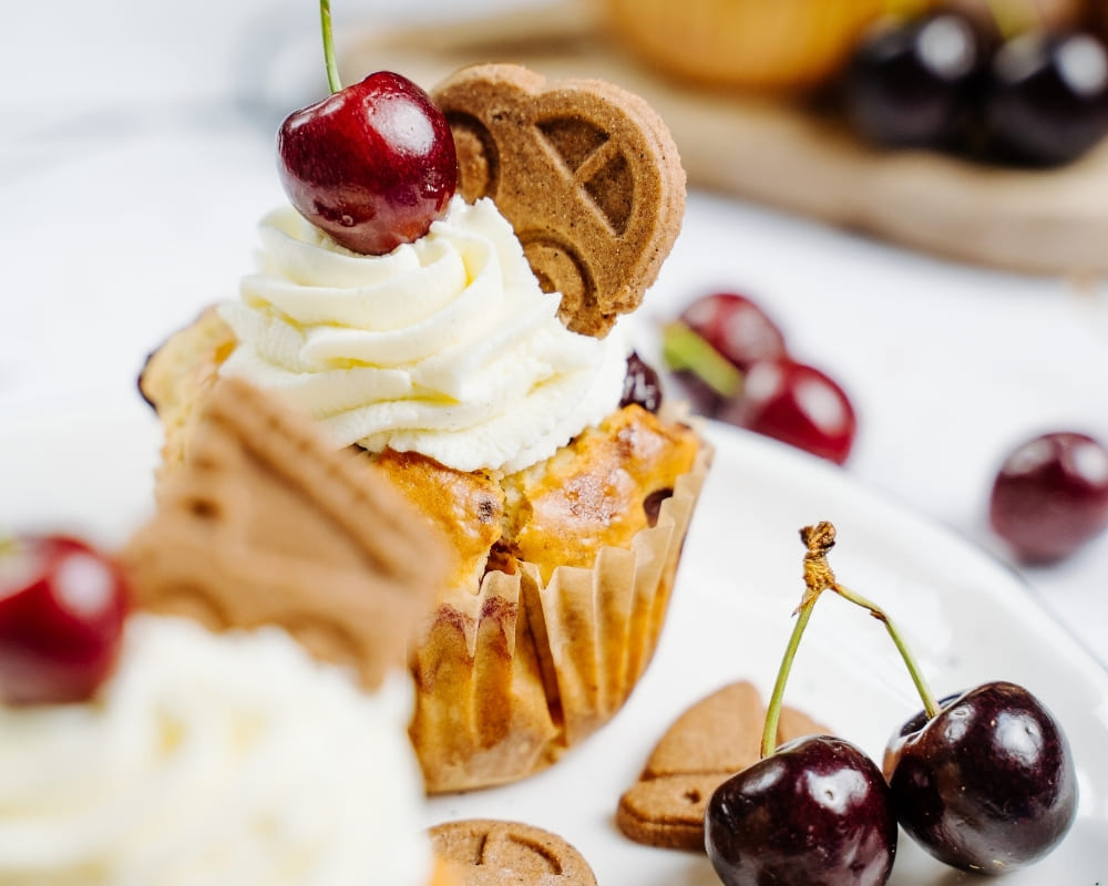 Bohlsener Mühle Muffin mit Sahnehaube, frischer Kirsche und einem knusprigen Cookie auf einem weißen Teller serviert.