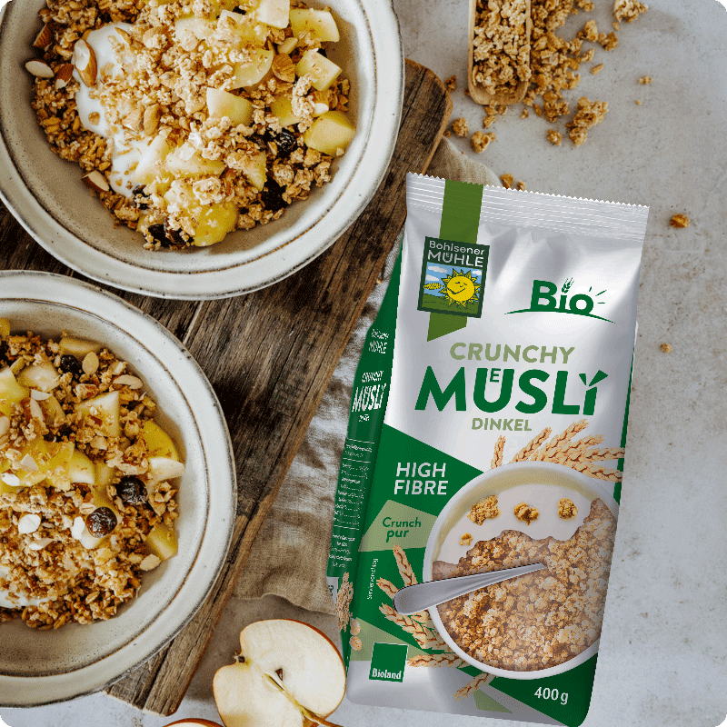 Bohlsener Mühle Dinkel Crunchy Müsli neben zwei Schalen mit Müsli, Joghurt und frischen Apfelstücken auf einem Holztisch.