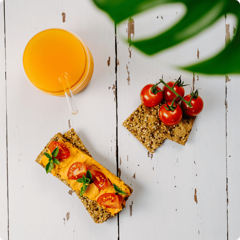 Bohlsener Mühle Knusper-Brot belegt mit Tomaten und Aufstrich neben einem Glas Saft auf weißem Holztisch.