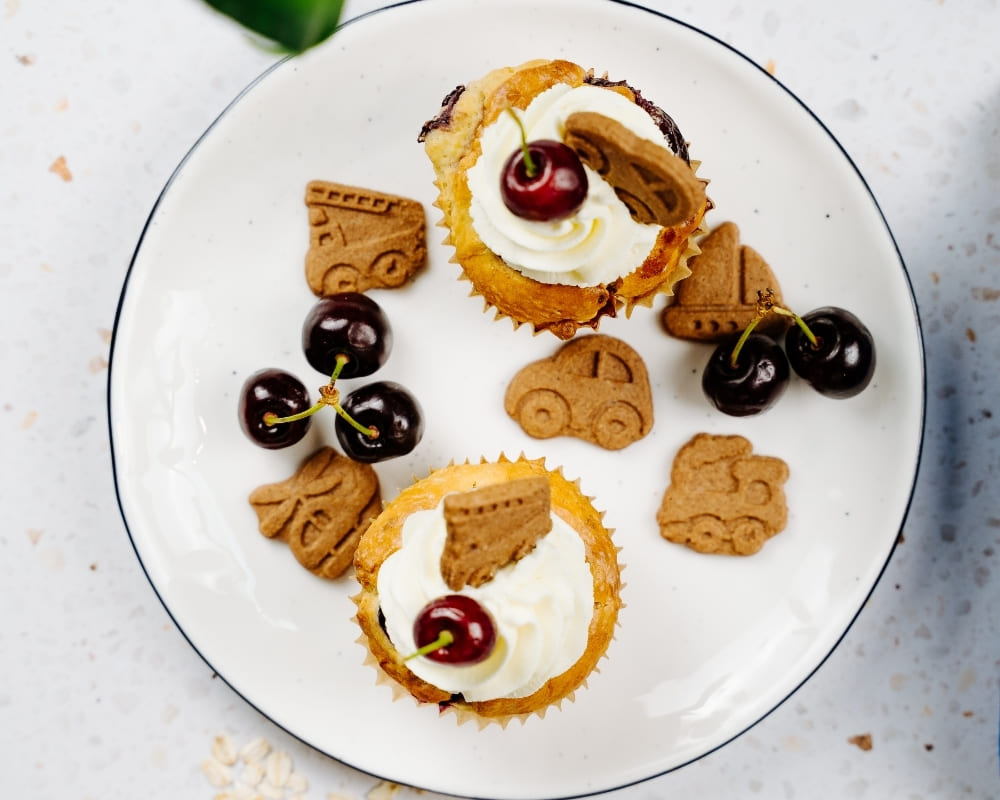 Zwei Bohlsener Mühle Muffins mit Sahne, Kirschen und Crunchy Keks-Figuren auf einem weißen Teller angerichtet.