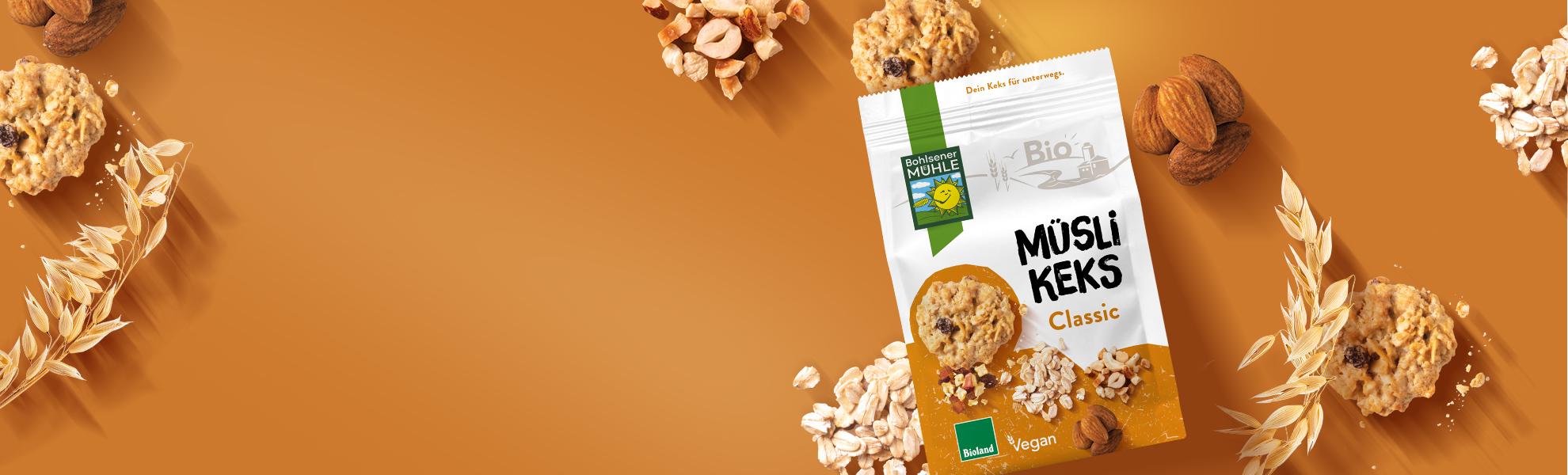 Bohlsener Mühle Müsli Keks Classic Packung umgeben von Haferflocken, Nüssen und knusprigen Cookies auf braunem Grund.