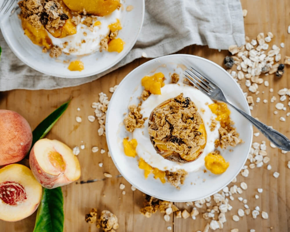 Zwei Teller mit gebackenen Pfirsichen mit Müsli und Joghurt, umgeben von frischen Pfirsichen und verstreuten Haferflocken auf einem Holztisch.