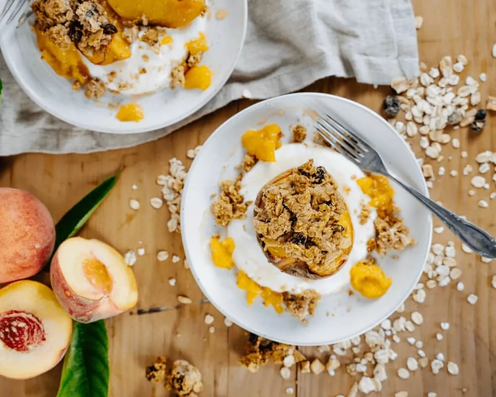 Zwei Teller mit gebackenen Pfirsichen mit Müsli und Joghurt, umgeben von frischen Pfirsichen und verstreuten Haferflocken auf einem Holztisch.