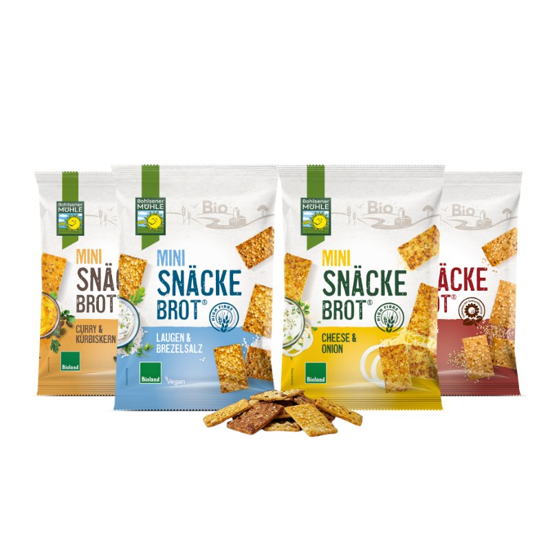 Vier Packungen Bohlsener Mühle Mini Snäcke Brot mit verschiedenen Sorten und losen Crunchy Stücken davor.