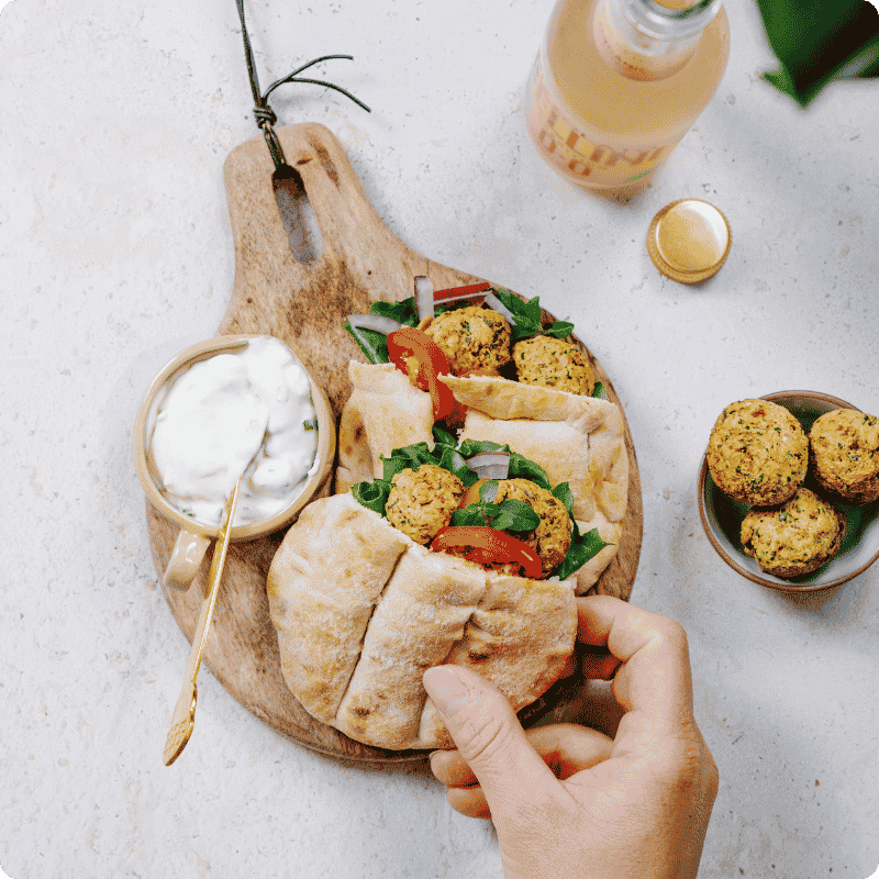 Eine Hand hält ein mit Bohlsener Mühle Falafel gefülltes Pitabrot auf einem Holzbrett neben einem Dip.