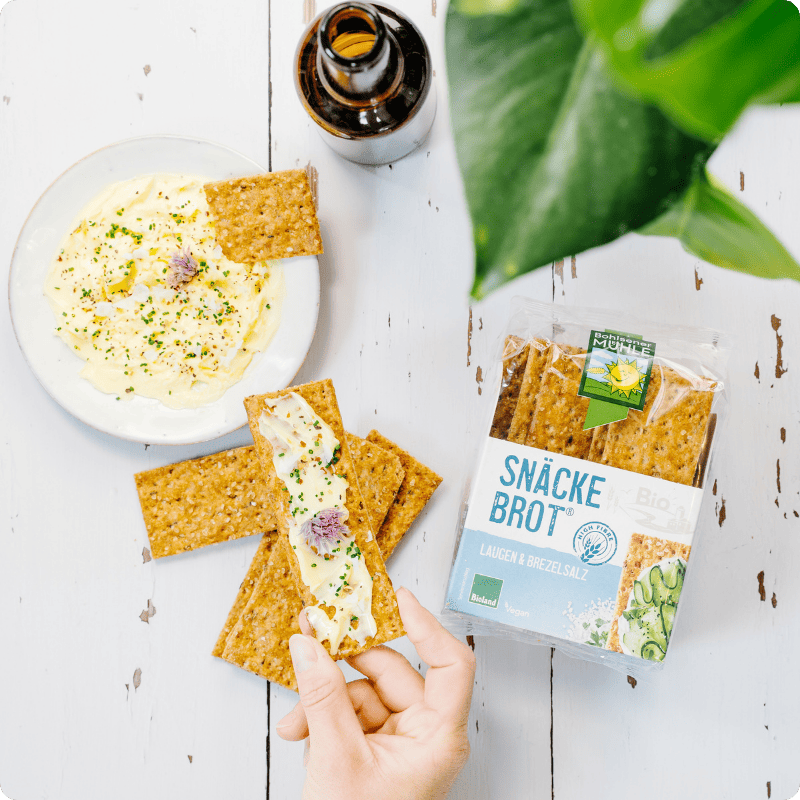 Eine Hand hält ein bestrichenes Bohlsener Mühle Snäcke Brot Laugen & Brezelsalz neben einer Schale mit Kräuterquark.