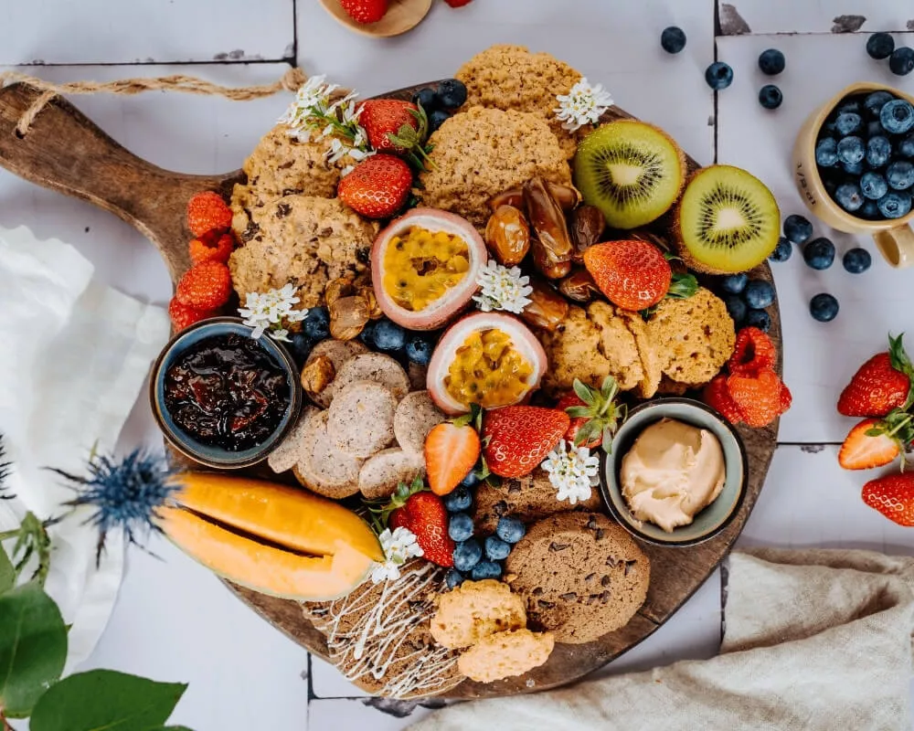 Ein Holzbrett gefüllt mit Bio-Keksen, Müsli, knusprigen Snacks und frischen Früchten der Bohlsener Mühle sowie frischen Früchten wie Erdbeeren, Kiwis, Blaubeeren und Passionsfrüchten, die nachhaltige Landwirtschaft und Artenvielfalt fördern.