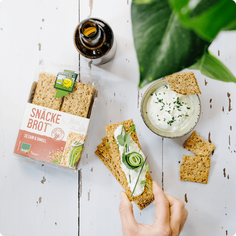Bohlsener Mühle Snäcke Brot Sesam & Dinkel mit Kräuterquark und Gurke auf einem hellen Holztisch serviert.