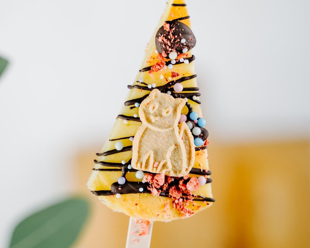 Ein festliches, dreieckiges Dessert am Stiel, dekoriert mit Schokoladenstreuseln, bunten Streuseln und einem Keks in Katzenform. Bohlsener Mühle.