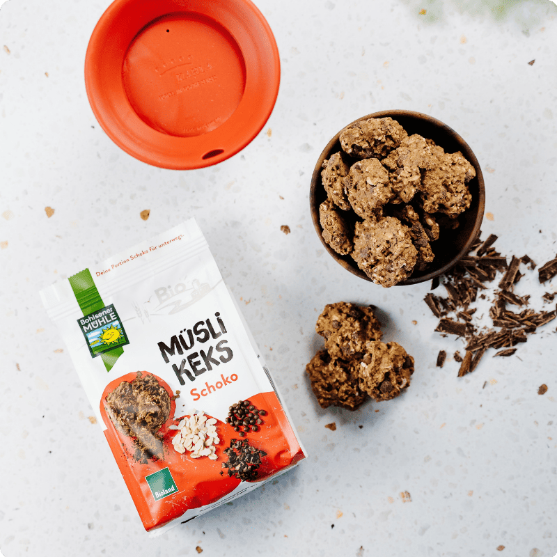 Bohlsener Mühle Müsli Keks Schoko in einer Schale neben der Packung und verstreuten Schokostückchen auf dem Tisch.