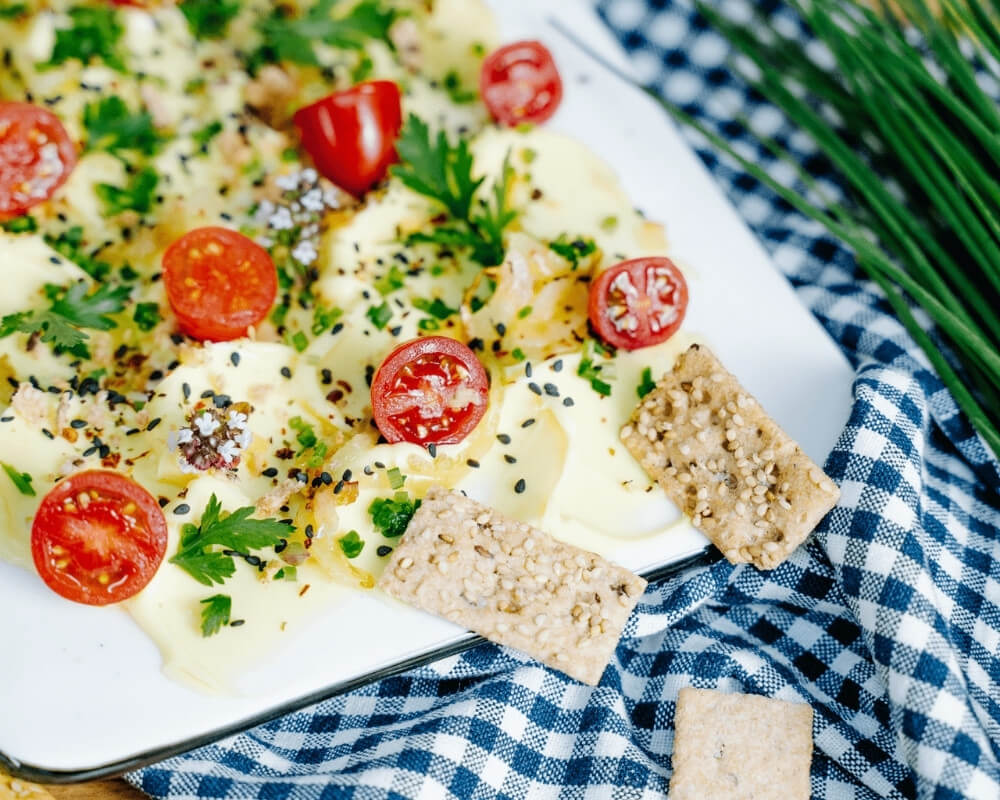Bio-Bohlsener-Mühle-Snack mit Butter, Kirschtomaten, Kräutern und Sesamcrackern auf einem karierten Tuch.