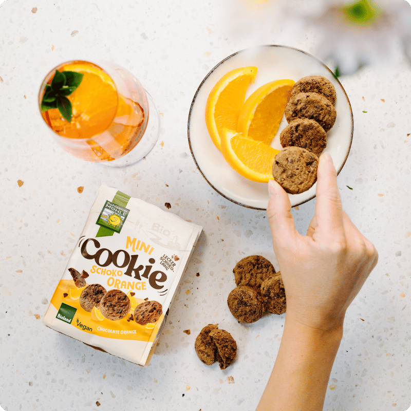 Eine Hand greift nach einem Mini Cookie Schoko Orange von Bohlsener Mühle neben einer Schale mit Orangen.