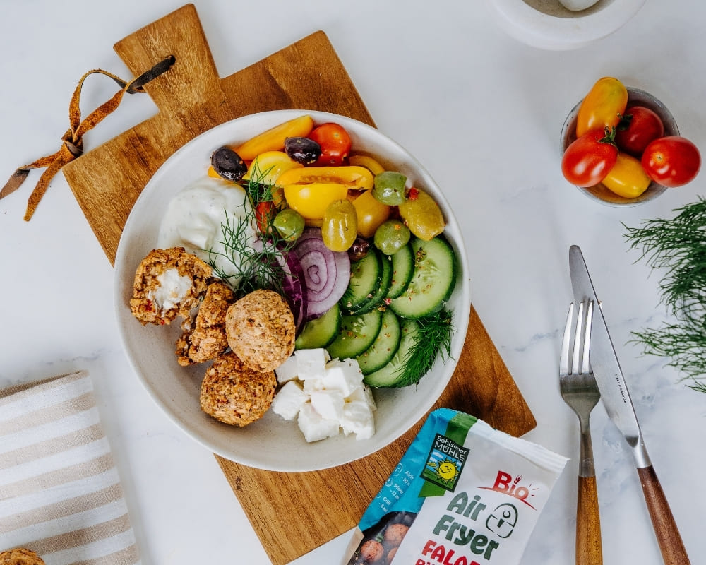 Bohlsener Mühle Air Fryer Falafel in einer Schüssel mit frischem Gemüse, Feta, Oliven und Dip auf einem Holzbrett.