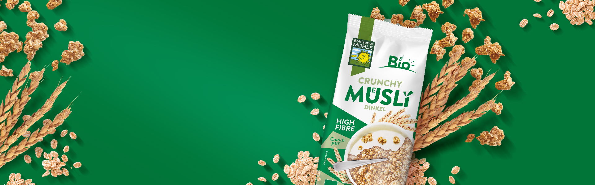 Bohlsener Mühle Crunchy Müsli Dinkel Packung umgeben von Getreideähren und Müslistücken auf grünem Hintergrund.